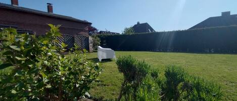 Ein grüner Hinterhof mit einem weißen Plastikstuhl, einem Ziegelhaus und einer Hecke.