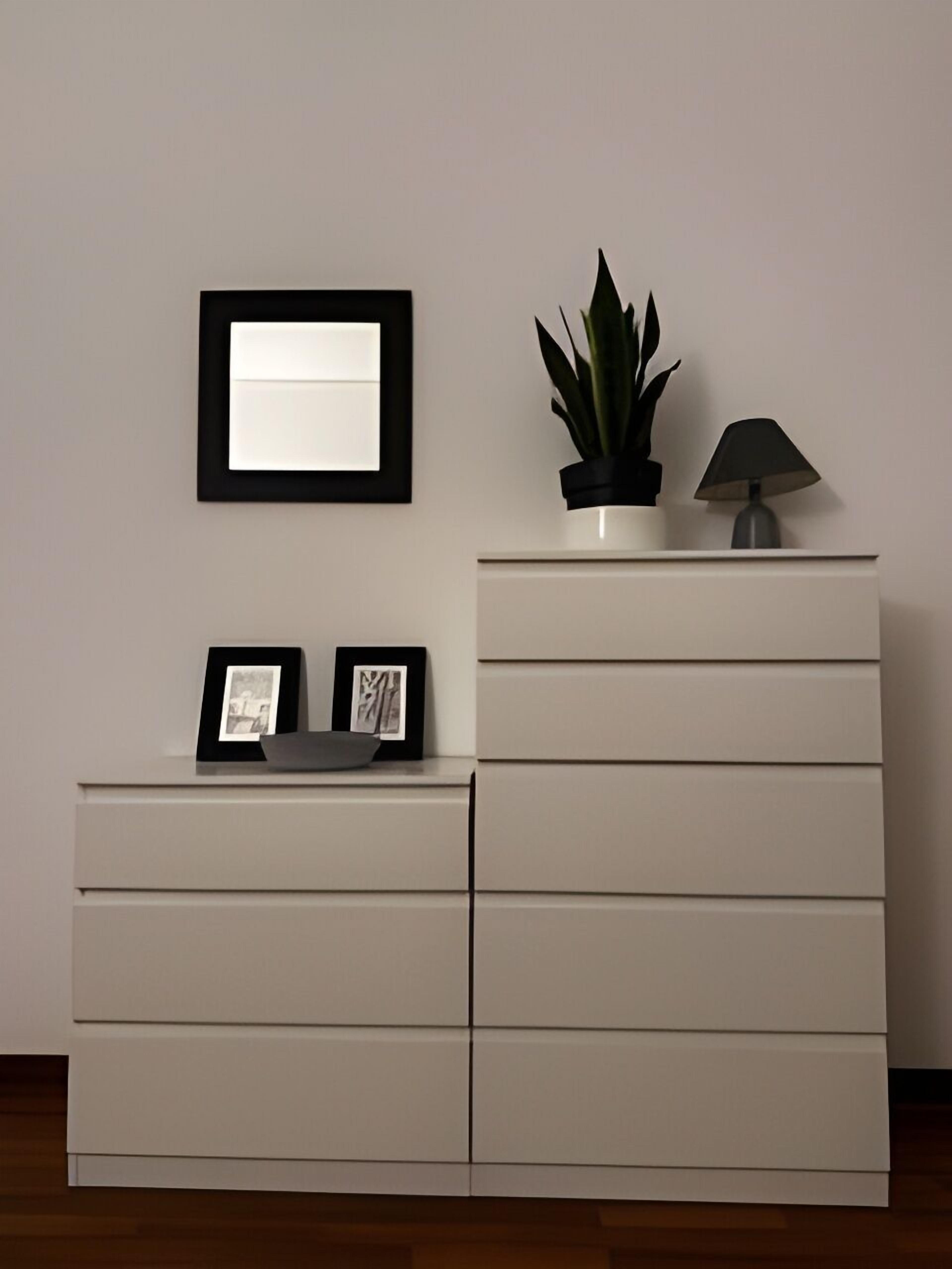 A white dresser with drawers, a framed picture, a potted plant, and a lamp on top.