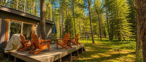 Une terrasse en bois avec des fauteuils Adirondack, un barbecue couvert et un chalet situé dans une forêt.