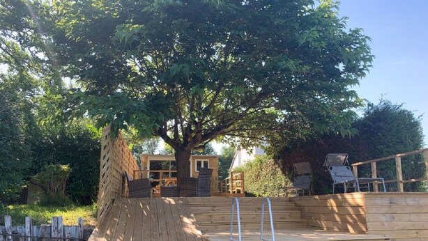 A wooden deck with a ladder leading to a platform, surrounded by trees and a fence.