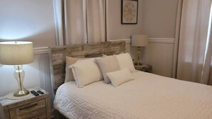 A neatly made bed with a white duvet and pillows, a wooden nightstand with a lamp and remote controls, and a framed picture on the wall. - Relaxing Poolside Condo (Savannah)