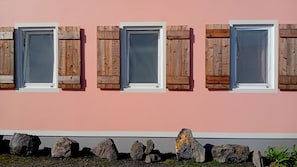 Eine Gebäudefassade mit drei Fenstern, jeweils mit Holzklapplädchen, vor einer rosafarbenen Wand.