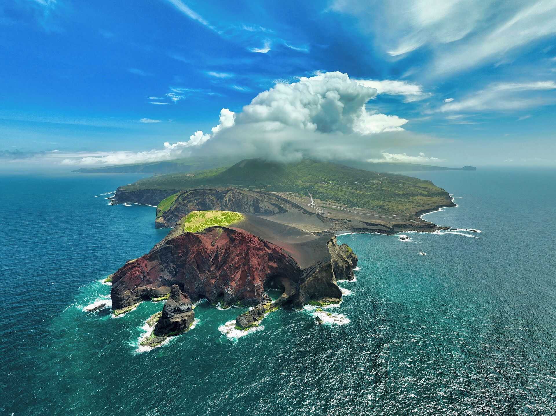 Vue aérienne d’une côte accidentée, avec un imposant sommet volcanique, un phare et un ciel d’un bleu limpide.