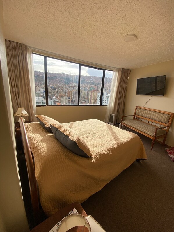 Une chambre d’hôtel avec un grand lit, un banc en bois, un téléviseur fixé au mur et une vue sur la ville par la fenêtre.
