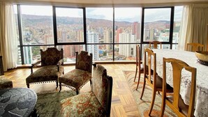 A room with a large city view through floor-to-ceiling windows, a dining area with wooden chairs and a table, and a patterned rug on the floor. - Central Apartment with Stunning Views in La Paz (La Paz)