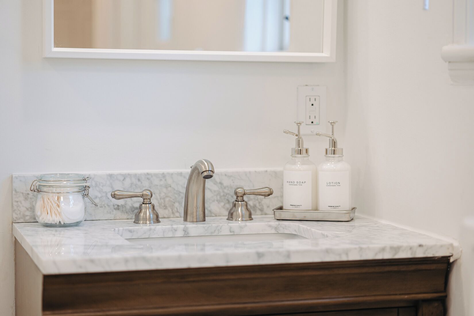 A bathroom with a marble sink, two faucets, a jar of cotton swabs, and two bottles of hand soap and lotion.