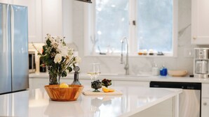 A modern kitchen with a central island, a vase with flowers, a wine glass, and a cutting board with fruits. - Charming Historic Home, Luxurious Remodel, Walk to Downtown, Tree-Lined Street (Delaware)