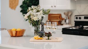 A kitchen counter with a wooden bowl of lemons, a vase with white flowers, a glass of white wine, and a decorative bottle. - Charming Historic Home, Luxurious Remodel, Walk to Downtown, Tree-Lined Street (Delaware)