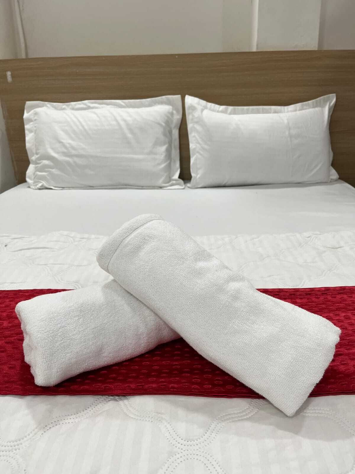 Un letto rifatto con biancheria e cuscini bianchi, una coperta rossa e un asciugamano bianco piegato.