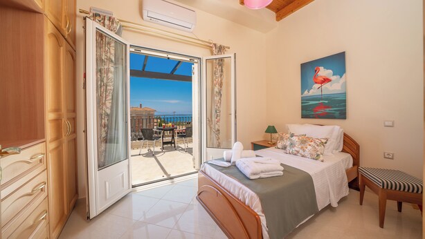 A bedroom with a bed, a bench, a painting, and an open door leading to a balcony with a view. - Irene Luxury Villa Residence | 10' drive to Lassi & Argostoli (Metaxata)