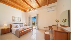 A bedroom with a wooden ceiling, a bed, a nightstand, a bedside table, a mirror, and a small table with a vase and flowers. - Irene Luxury Villa Residence | 10' drive to Lassi & Argostoli (Metaxata)