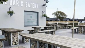 Ett utomhusområde med sittplatser, träbord och stenpelare, en skylt med texten ”BAR & RESTAURANG” och ett fönster med en logotyp.