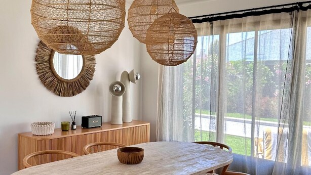 Un coin repas avec une table en bois, des suspensions en osier, un miroir et une vue sur la verdure à travers des rideaux transparents.