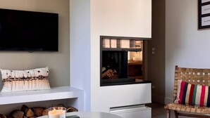 Un salon moderne avec un téléviseur à écran plat fixé au mur, une cheminée, un coin salon confortable avec un fauteuil en osier et une petite table sur laquelle est posée une bougie.