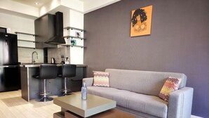 Ein modernes Wohnzimmer mit einem grauen Sofa, zwei schwarzen Barhocker, einem Couchtisch und einer Küchenzeile mit einer schwarzen Dunstabzugshaube und einer Mikrowelle.