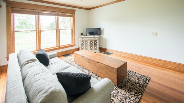 Ein Wohnzimmer mit einer Couch, einem Couchtisch, einem Fernseher auf einem Schrank und einem großen Fenster mit Jalousien.