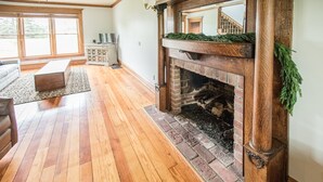 A spacious living room with hardwood flooring, a fireplace with a brick surround, and a large window with a view of the outdoors.