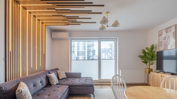 Un salon moderne comprenant un canapé d’angle, une table à manger avec des chaises, une grande fenêtre avec des stores et un téléviseur à écran plat posé sur un meuble en bois.