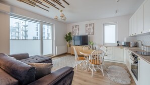Une cuisine moderne avec des meubles blancs, un coin repas avec une table et des chaises en bois, et une grande fenêtre donnant sur des immeubles.