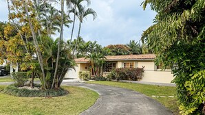 A single-story house with a red-tiled roof, surrounded by lush greenery and palm trees.