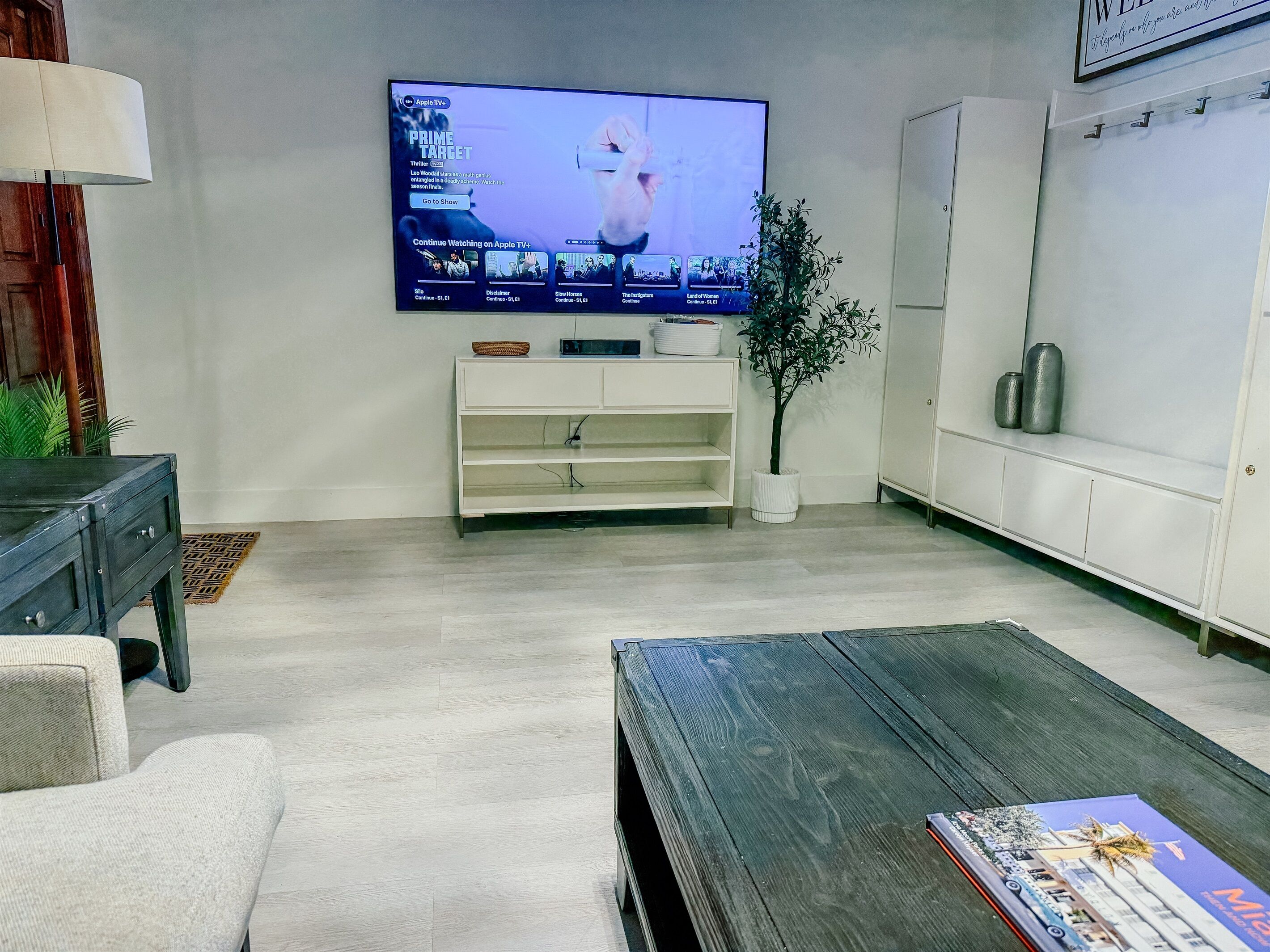 Ein modernes Wohnzimmer mit einem an der Wand befestigten Flachbildfernseher, einer weißen Unterhaltungseinheit darunter, einem Holztisch, einer Couch und einer Topfpflanze.