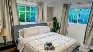 A neatly arranged bedroom with a bed, a nightstand, a lamp, and a view of greenery through the windows.