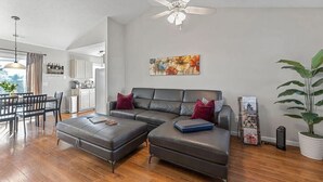 A modern living room with a sectional sofa, a dining area with a table and chairs, a ceiling fan, and a large window. - 4 Bed Beach Getaway! Fenced Yard + Garage Parking (Wilmington)