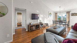 A modern living room with a flat-screen TV, a dining area with a table and chairs, and a large window with curtains. - 4 Bed Beach Getaway! Fenced Yard + Garage Parking (Wilmington)