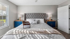 A bedroom with a tufted headboard, a bed with a patterned bedspread, two bedside tables with lamps, a window with blinds, and a door. - 4 Bed Beach Getaway! Fenced Yard + Garage Parking (Wilmington)
