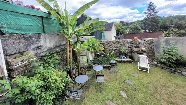Un patio trasero con muro de piedra, un árbol de plátano, una parrilla y una pequeña zona de estar con dos sillas y una mesa.