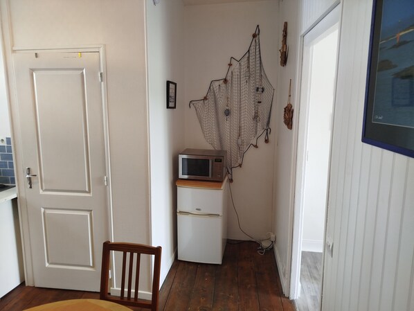 Una habitación con piso de madera, una puerta y un refrigerador blancos, un microondas encima del refrigerador y una silla.