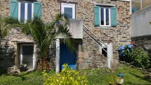 Un edificio de piedra con puerta azul y contraventanas verdes, una escalera y un jardín con plantas en macetas.