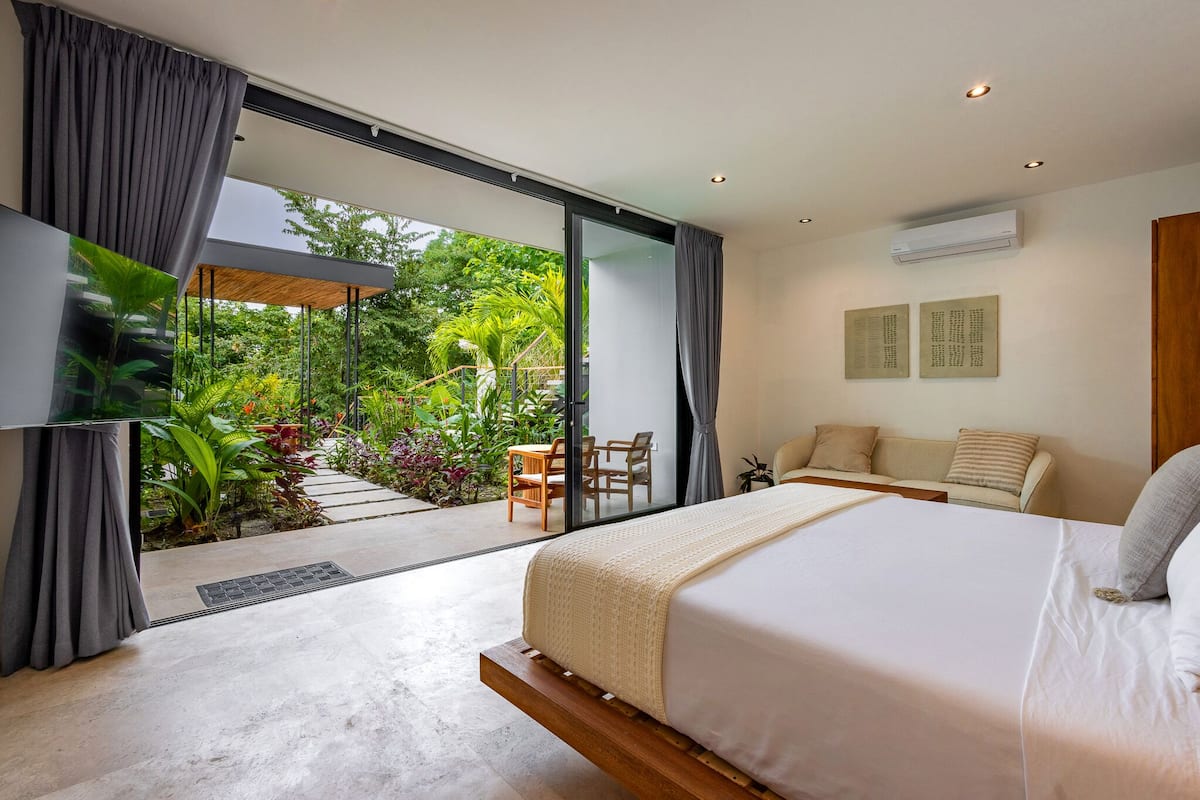 A modern bedroom with a large bed, a TV, and a view of a garden through a sliding glass door.