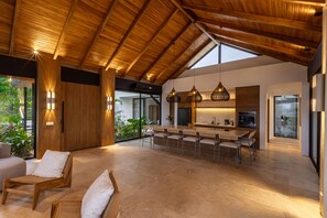 A spacious kitchen with a high ceiling and wooden beams, a dining area with chairs and a table, and a view of greenery outside. - Bahia Rooms (Cóbano)