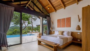 Un dormitorio con una cama grande, cabecera de madera y un banco. Cuenta con un ventanal con cortinas, vista a la vegetación y una piscina exterior.