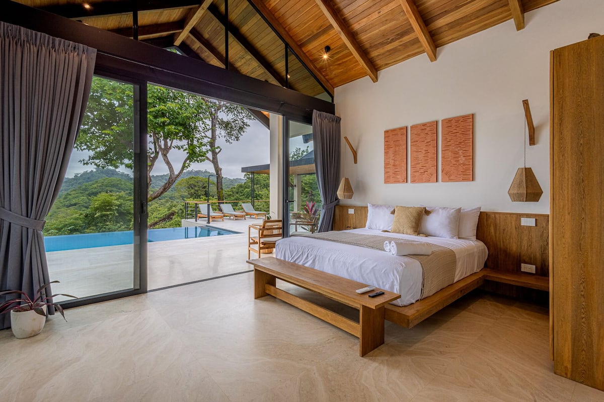 A bedroom with a large bed, wooden headboard, and a bench. It features a large window with curtains, a view of greenery, and a pool outside.