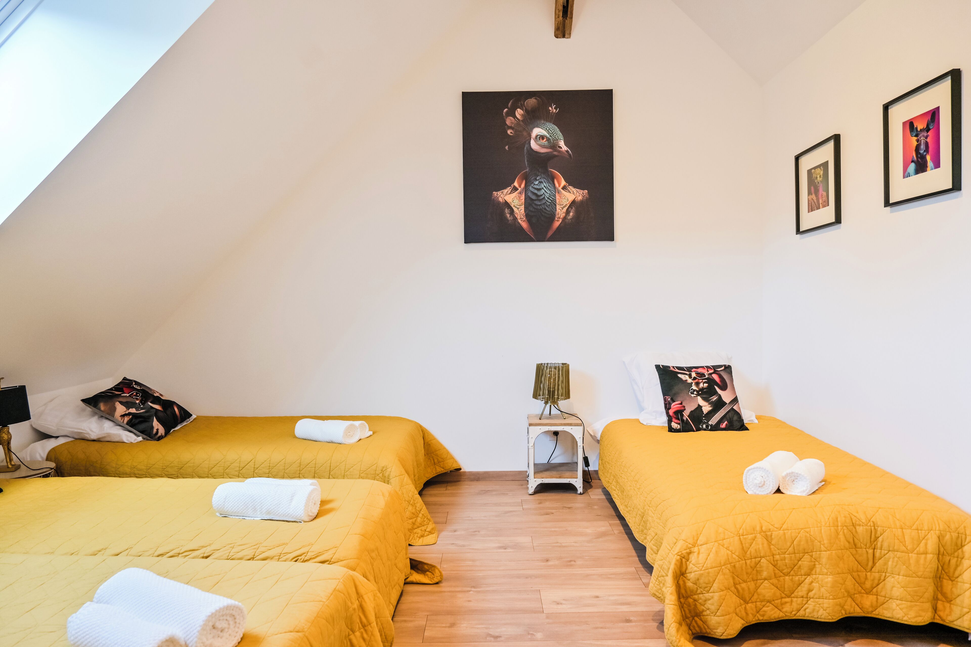 Habitación con dos camas, cada una con una colcha amarilla y toallas blancas. Se aprecia un cuadro en la pared y una mesita de noche con una lámpara.