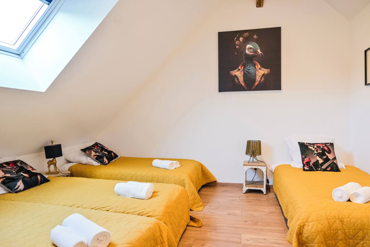 Une chambre d’hôtel avec deux lits, chacun recouvert d’une couvre-lit jaune et agrémenté d’oreillers assortis. Un tableau est accroché au mur au-dessus des lits, il y a une petite table avec une lampe, et une fenêtre d’où l’on peut admirer le ciel.