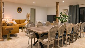 A dining area with a wooden table and wicker chairs, a sofa with cushions, and a television on the wall. - Les Longères de Montfranc (Pierrefitte-sur-Sauldre)