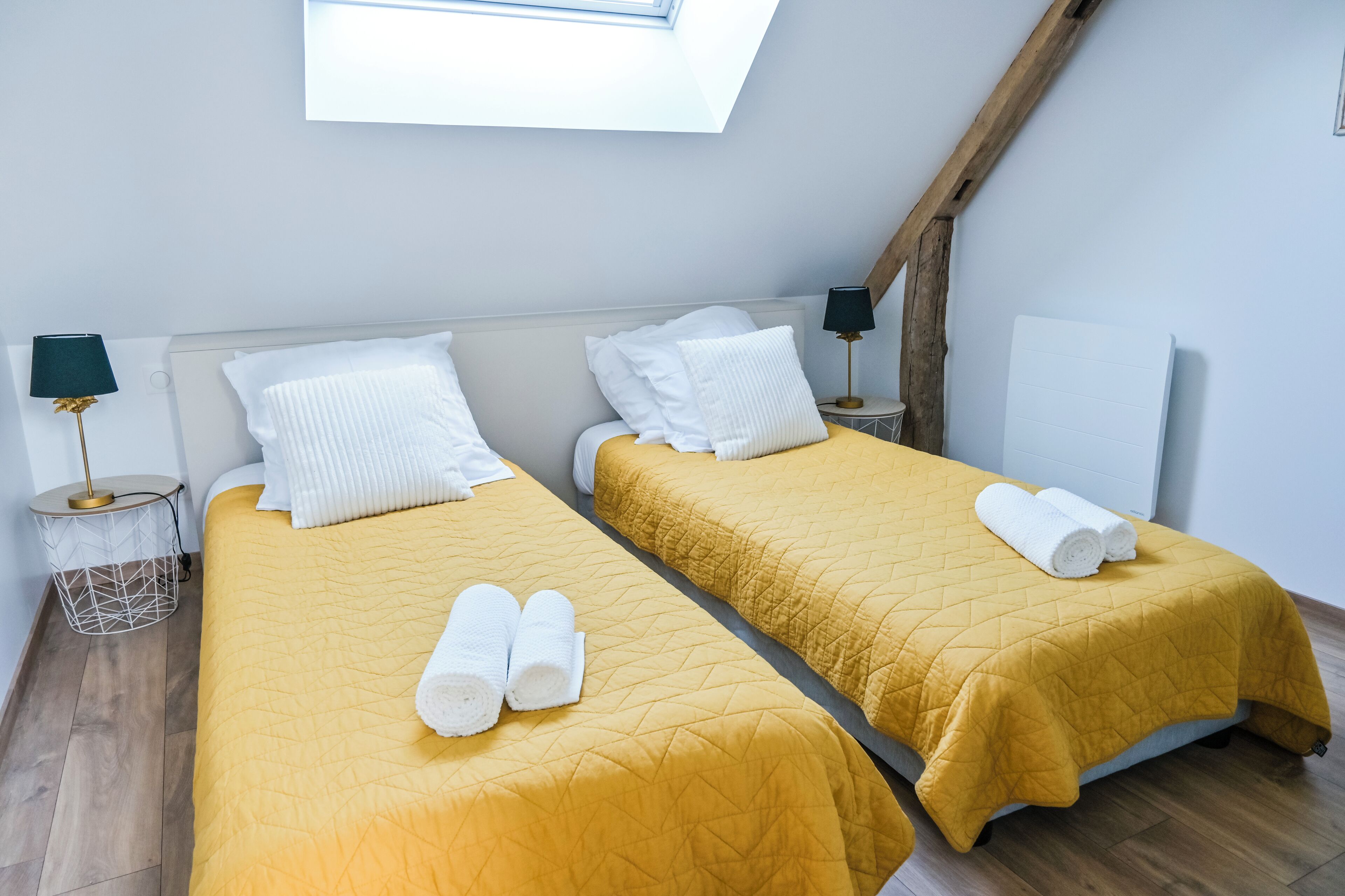 Habitación con dos camas, cada una con colcha amarilla acolchada y almohadas blancas. Hay toallas enrolladas sobre las camas y un tragaluz arriba.