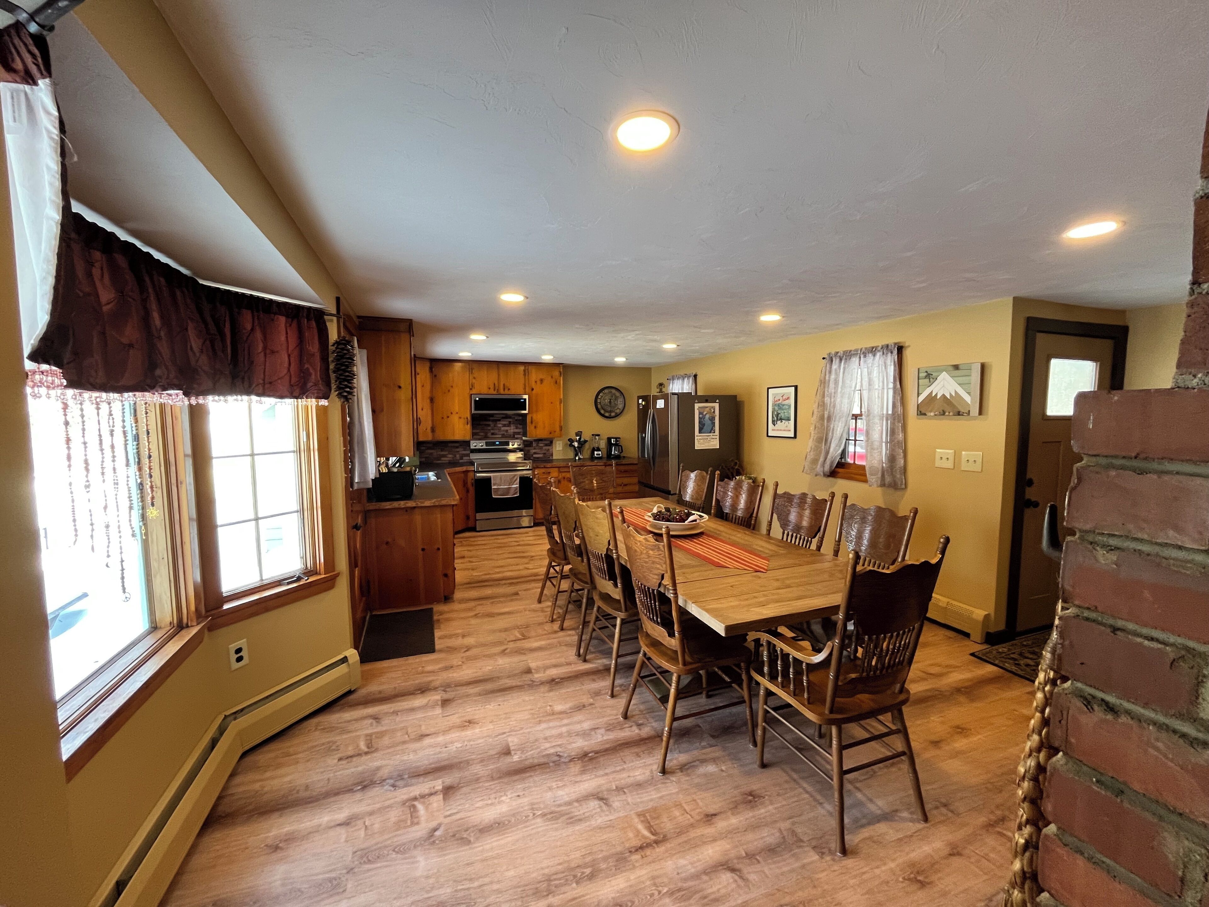 A dining area with a wooden table and chairs, a kitchen in the background, and a brick wall.