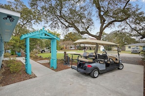 Un golf cart parcheggiato di fronte a un arco turchese, un edificio con tetto bianco e un grande albero sullo sfondo.