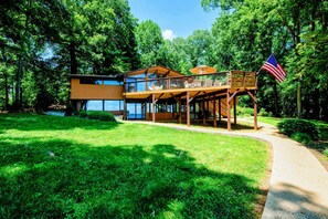 A modern two-story house with a deck, surrounded by lush greenery and a clear sky.