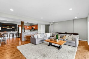 Een moderne woonkamer met een grijze bank, een houten salontafel en een keuken op de achtergrond.