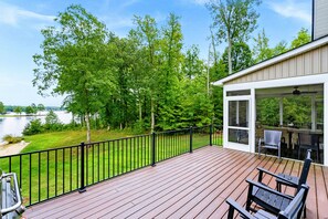 Une terrasse en bois offrant une vue sur un lac et des arbres, un espace extérieur couvert avec des chaises et des tables, et un bâtiment doté de grandes fenêtres.