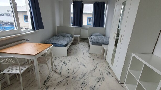 A compact room with a bed, a desk, and a chair. There are windows with curtains, a radiator, and a wardrobe. - Akdeniz Pension (Ludwigsfelde)