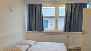 A bedroom with a bed, a bedside table, a radiator, and a window with curtains. - Akdeniz Pension (Ludwigsfelde)