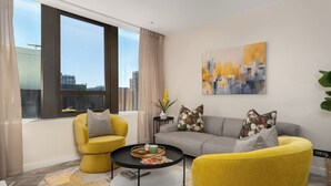 A modern living room with a sofa, two yellow armchairs, a coffee table, and a large window with a city view. - Designer One Bed Apt | No Loadshedding (Cape Town)