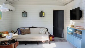 A room with a white wooden wall, a sofa with cushions, a small table with a plant, a clock, and a television on a blue cabinet. - New listing! Cozy, fresh and clean 1-bedroom cottage with Pond access in Chatham (Chatham)