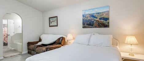 A bedroom with a large bed, two bedside lamps, a ceiling fan, a framed picture, and a door leading to another room.